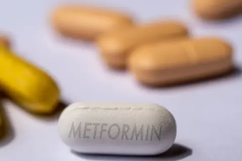 Eine weiße Tablette mit der Aufschrift "Metformin" liegt im Vordergrund. Im Hintergrund sind unscharfe gelbe und orangefarbene Pillen zu sehen. Forschung untersucht, ob Metformin bei Demenz helfen kann.