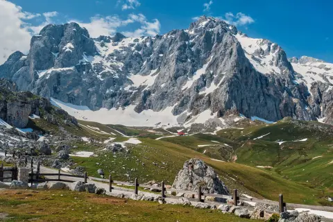 Majestuosas montañas nevadas en los Picos de Europa, cerca de Llanes. Un camino de piedra y madera serpentea por prados verdes salpicados de nieve.