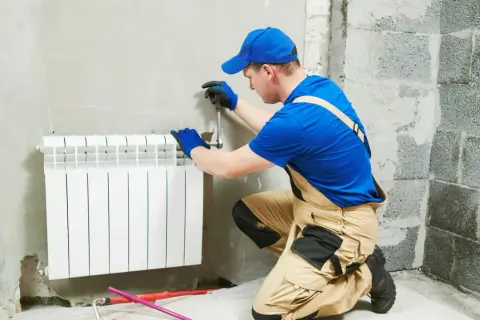 Instalator w niebieskiej czapce i ogrodniczkach pracuje przy grzejniku. Pokazuje, jak zdjąć grzejnik, używając klucza.