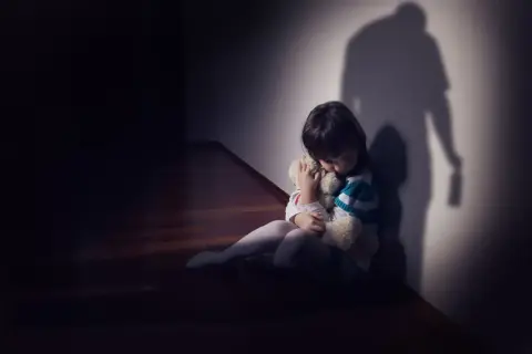 Dziecko tuli misia, skulone w kącie. Cień dorosłego z butelką sugeruje agresję i strach.