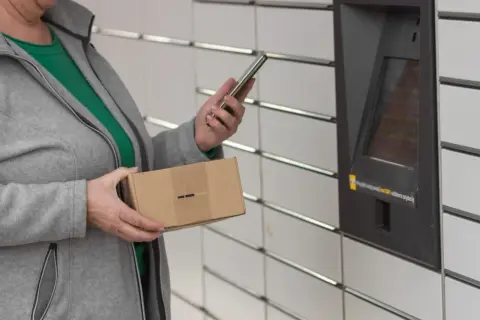 Kobieta z paczką w ręku korzysta z telefonu przy automacie InPost. Szybkie przekierowanie paczki jest teraz łatwiejsze.