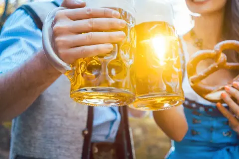Zwei Hände halten je ein **Mass Bier** und stoßen an. Eine Brezel liegt in der anderen Hand.