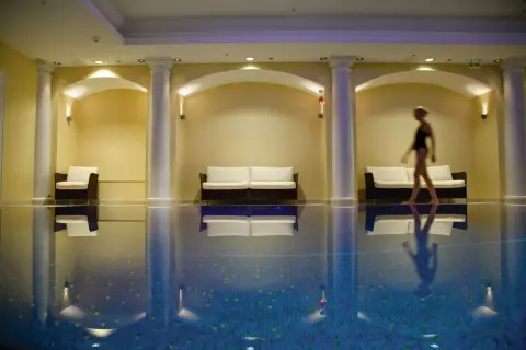 Una mujer camina junto a una piscina interior, rodeada de sofás y columnas, creando una atmósfera de relajación para una experiencia spa sensorial.