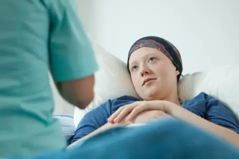 Młoda pacjentka w chustce na głowie leży w łóżku, słuchając pielęgniarki. To może być początek walki z chorobą, gdzie rak przedinwazyjny co to znaczy, jest kluczowym pytaniem.