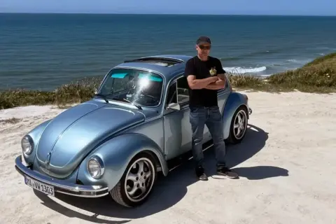 Man står bredvid en ljusblå Volkswagen Bubbla vid havet. En påminnelse om vem grundade Volkswagen och dess ikoniska design.