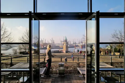 Blick durch offene Glastüren auf den Hamburger Hafen mit dem alten Elbtor und Kränen. Eine Frau genießt die Aussicht, perfekt für einen Stintfang Hamburg.