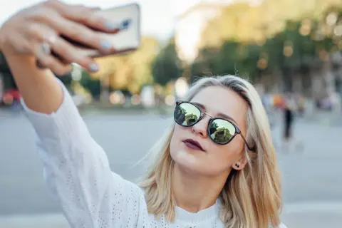 Blondynka w okularach robi sobie selfie telefonem. W okularach odbija się budynek. Oto jak zrobić dobre selfie telefonem.