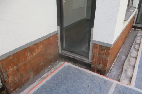 Rote Ziegelsteine am Sockel zeigen Anzeichen von aufsteigender Feuchtigkeit im Sockelbereich. Ein Fensterrahmen ist sichtbar.