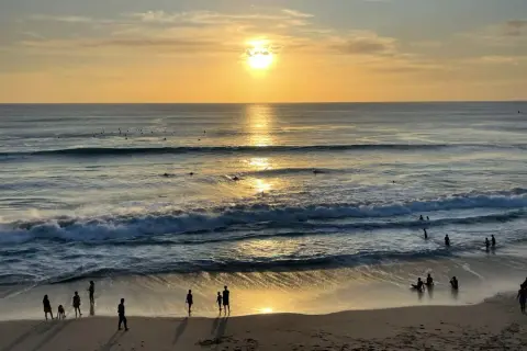 Zachód słońca na plażach Bali. Surferzy czekają na fale, a ludzie spacerują po piasku, ciesząc się złotym blaskiem.