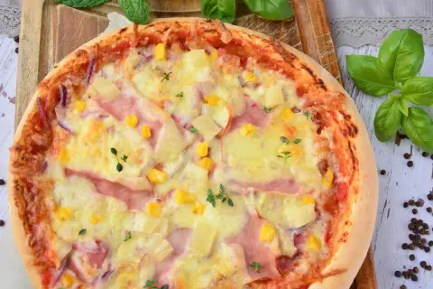 Apetyczna pizza z ananasem, szynką, kukurydzą i serem, ozdobiona świeżymi ziołami.