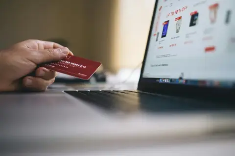 Mano sosteniendo tarjeta roja para pagar en plataformas de compra online. Pantalla de portátil muestra ofertas.