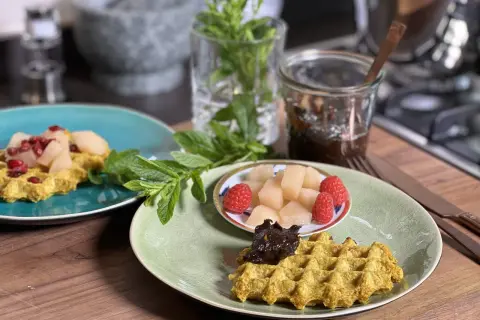 Zwei Teller mit goldgelben Haferflocken Waffeln, garniert mit Früchten und Marmelade, daneben frische Minze.