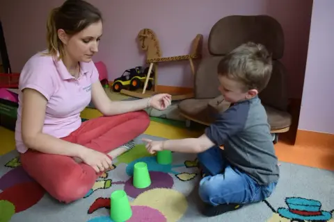 Terapia behawioralna autyzm ćwiczenia: chłopiec i terapeutka bawią się kubeczkami na dywanie.