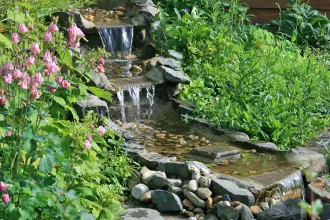 Ein kleiner Wasserfall im Garten, umgeben von grünen Pflanzen und rosa Blüten. Tolle Ideen für einen naturnahen Garten.