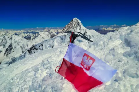 Na śnieżnym szczycie, z lodową siekierą i polską flagą, rozpościera się widok na skalisty, szeroki szczyt i góry.