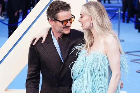 Pedro Pascal y su pareja, con ella luciendo un vestido azul con flecos, se miran tiernamente en una alfombra azul.