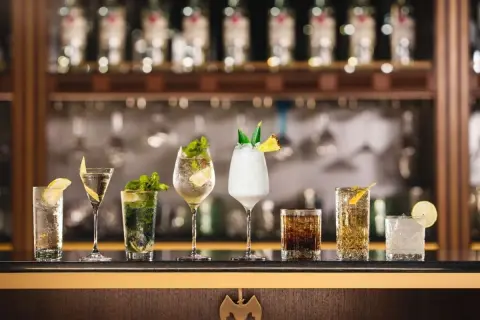 A row of seven colorful cocktails on a bar, with bottles in the background.