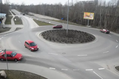 Widok z góry na rondo z kilkoma samochodami, drzewami i billboardami w tle.