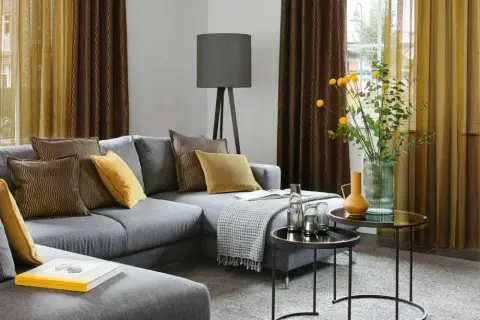 Gemütliches Wohnzimmer mit grauem Sofa, Kissen in Braun und Gelb, sowie moderne Fenstergestaltung mit zweifarbigen Vorhängen.