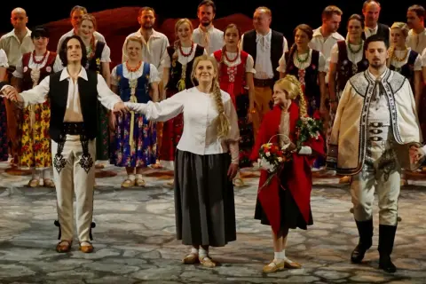 Grupa ludzi w strojach ludowych, mężczyzna w białej koszuli, kamizelce i spodniach z haftem, kobieta w kwiecistej sukni i halka.
