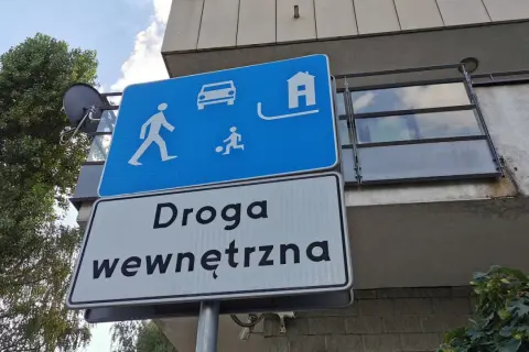 Znak "strefa zamieszkania" informuje o ograniczeniu prędkości. Widoczni piesi, dziecko grające w piłkę, samochód i dom.