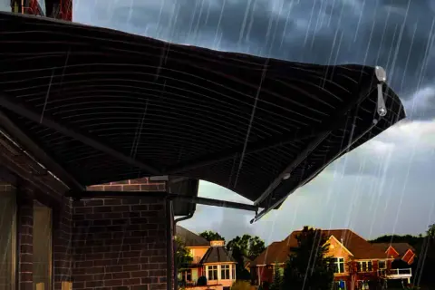 A dark, striped awning installation provides shelter from a heavy rain shower over a brick house.