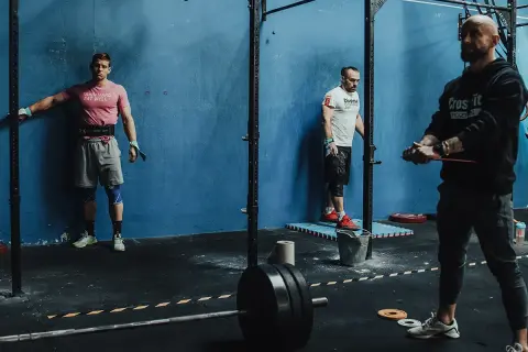 Trzech mężczyzn w siłowni CrossFit, jeden oparty o ścianę, drugi przy stojaku, trzeci z gumą treningową.