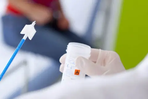 Ręka w rękawiczce trzyma pojemnik na próbkę do cytologii płynnej. W tle widać pacjentkę.