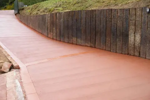 Camino de hormigón cepillado con borde de ladrillo, flanqueado por un muro de traviesas de madera.