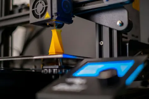 Drukarka 3D drukuje żółty obiekt, zbliżenie na dyszę i platformę roboczą.