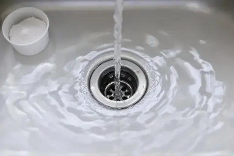 Eau courante dans un évier, avec une tasse de bicarbonate de soude à côté, pour déboucher le lavabo.