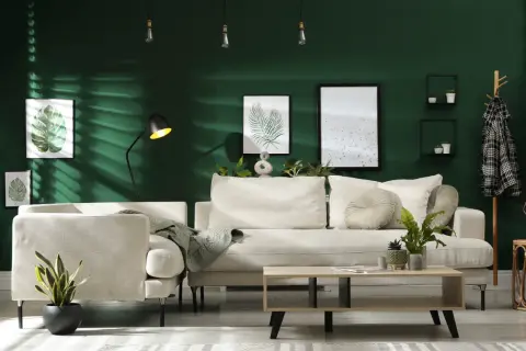Ein gemütliches Wohnzimmer mit beiger Couch, Kaffeetisch und Pflanzen vor einer dunkelgrünen Wand. Ein Kleiderständer mit kariertem Mantel und ein trendiger Wandbehang vervollständigen das Bild.