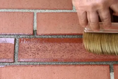 Mano con pennello applica prodotto per restauro mattoni faccia a vista, rinnovando la parete.