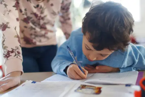 Chłopiec pisze w zeszycie, a obok niego stoi osoba dorosła.