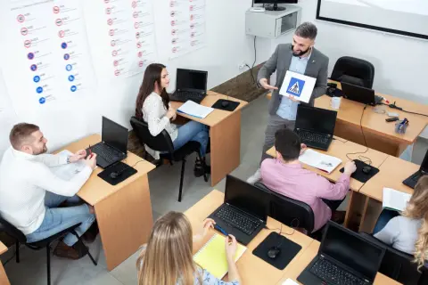 Nauczyciel pokazuje znak przejścia dla pieszych, omawiając zasady ruchu drogowego i kasowanie punktów karnych. Uczniowie słuchają, pracując przy laptopach.