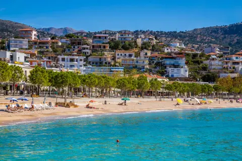 Piękna, słoneczna **Albania** w **czerwcu**. Ludzie odpoczywają na plaży, woda jest turkusowa, a miasteczko malowniczo wznosi się na wzgórzu.