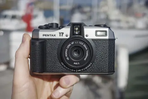A hand holds the highest quality film camera, a silver and black Pentax 17, with a blurred marina in the background.