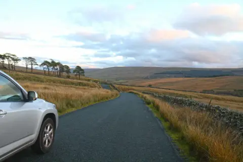 Voiture argentée sur une route sinueuse dans un paysage vallonné. La conduite à gauche est visible sur le côté de la route.