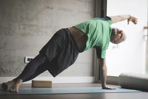 Hombre mayor con barba gris practica yoga para hombres en una esterilla, en postura de plancha lateral.