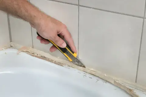 Ręka z nożykiem usuwa stary silikon z brodzika. Dowiedz się, czym usunąć silikon z brodzika, by przygotować go do nowej fugi.