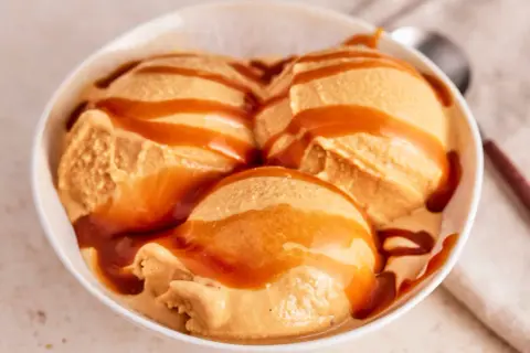 Drei Kugeln cremiges Karamell-Eis, übergossen mit flüssigem Karamell, in einer weißen Schüssel.