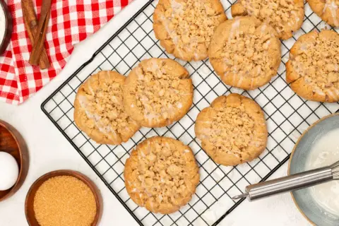 Frisch gebackene Zimt-Streusel-Kekse auf einem Kuchengitter. Zutaten wie Eier, Zucker und Mehl sind daneben zu sehen.
