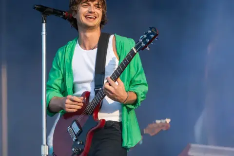 Joe Keery w zielonej koszuli i białej podkoszulce gra na czerwonej gitarze elektrycznej na koncercie.