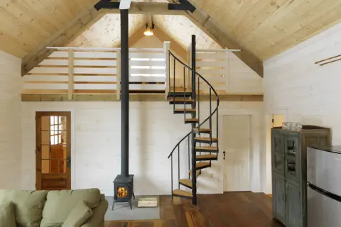 Idee soppalco basso: scala a chiocciola in metallo e legno porta a un soppalco con ringhiera in legno, stufa a legna e divano.