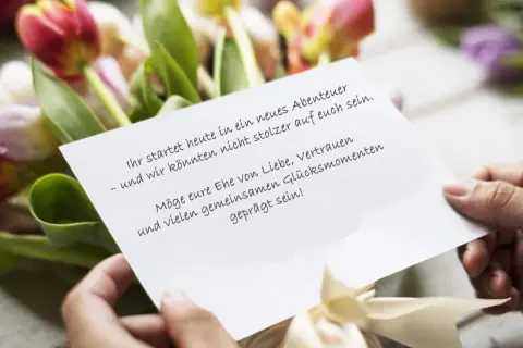 Hände halten eine Karte mit Hochzeitsglückwünschen vor Tulpen. "Ihr startet heute in ein neues Abenteuer – und wir könnten nicht stolzer auf euch sein. Möge eure Ehe von Liebe, Vertrauen und vielen gemeinsamen Glücksmomenten geprägt sein!