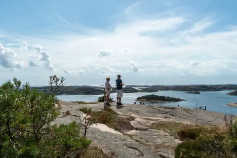 Två personer njuter av utsikten från Vetteberget i Fjällbacka. Vackert väder med moln och sol över skärgården.