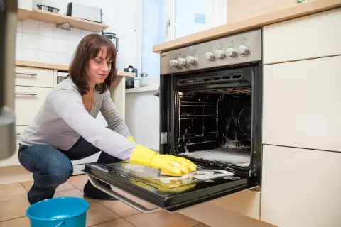 Frau mit gelben Handschuhen reinigt den Backofen. Schaum und ein blauer Eimer stehen bereit.