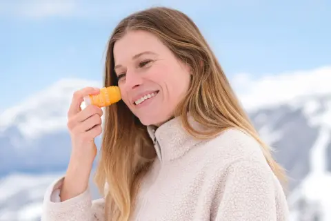 En hiver, faut-il mettre de la crème solaire ? Cette femme souriante en applique sur son visage, profitant d'un paysage enneigé.