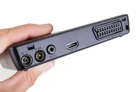 Dłoń trzyma czarny dekoder z portami RF IN, RF OUT, COAXIAL, HDMI OUT i SCART.