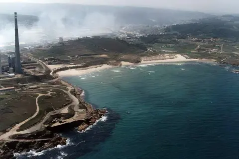 Vista aérea de la playa de Sabón, con una chimenea industrial y humo en el fondo, y el mar azul turquesa.
